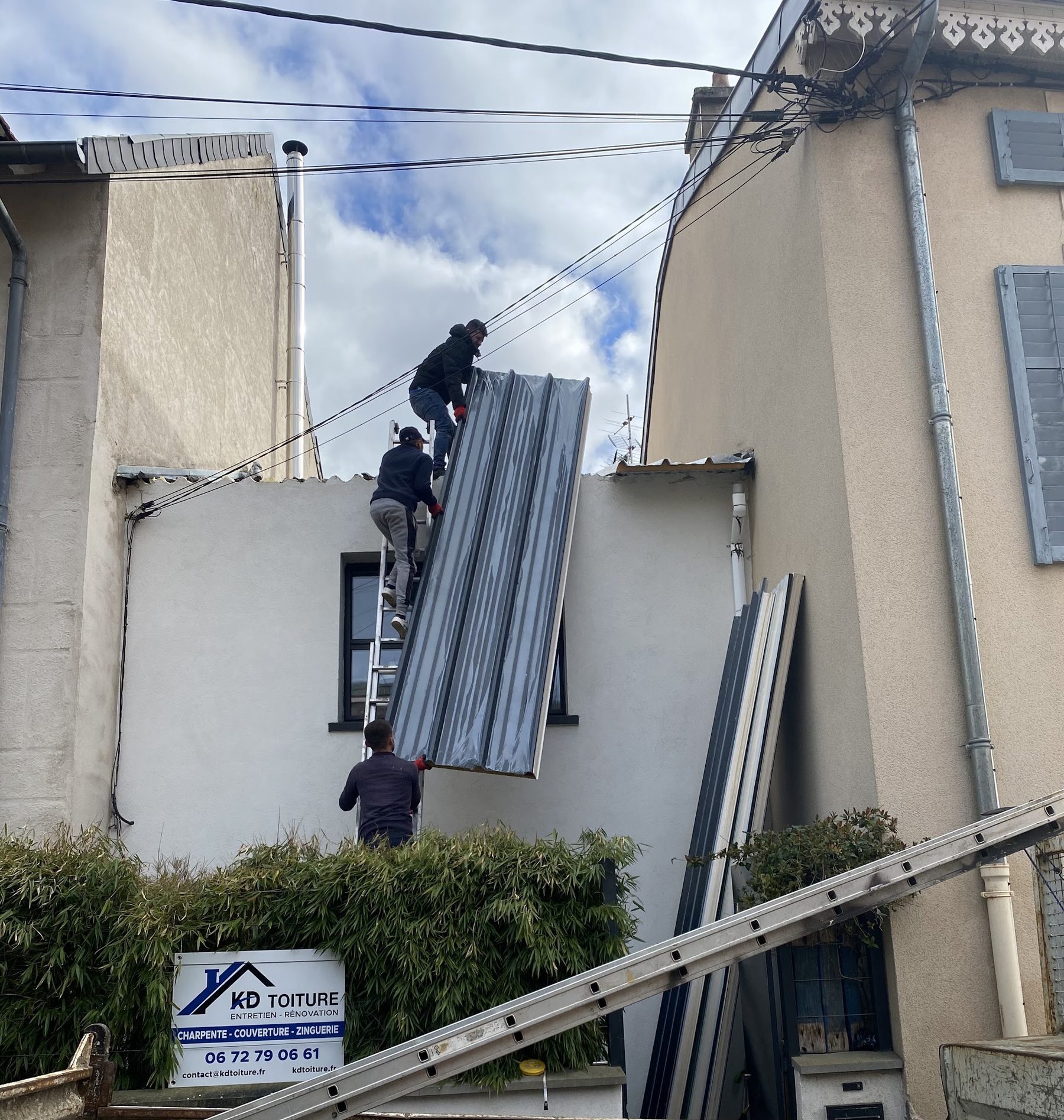 Trois couvreurs KD Toiture montent un panneau à l'échelle, le panneau de chantier KD au pied du mur